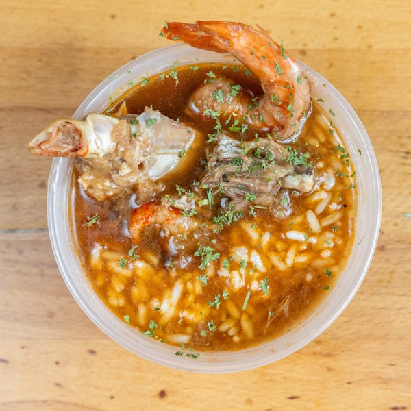 Top-down view of a seafood soup with shrimp, fish, vegetables, rice, and chopped herbs in a plastic bowl.