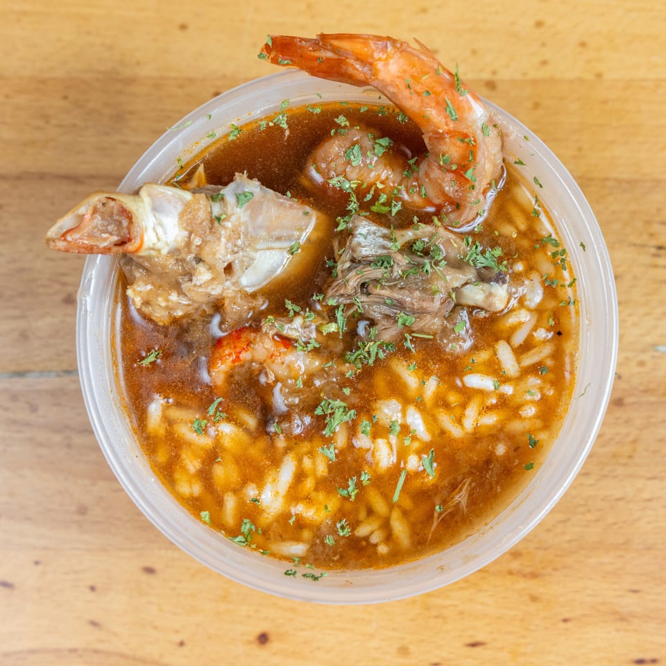 Top-down view of a seafood soup with shrimp, fish, vegetables, rice, and chopped herbs in a plastic bowl.