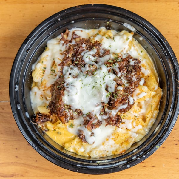 Cheesy pasta topped with shredded roasted meat and herbs in a black bowl on a wooden table.