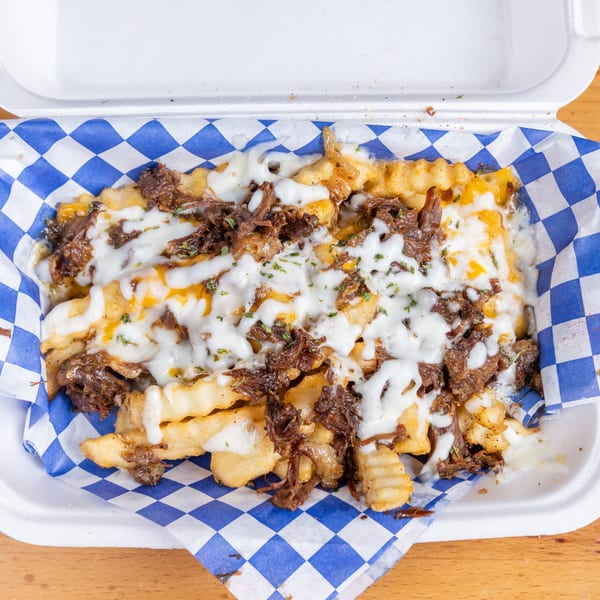 Loaded fries topped with shredded beef, melted white and cheddar cheese, and herbs in a checkered-lined takeout container.