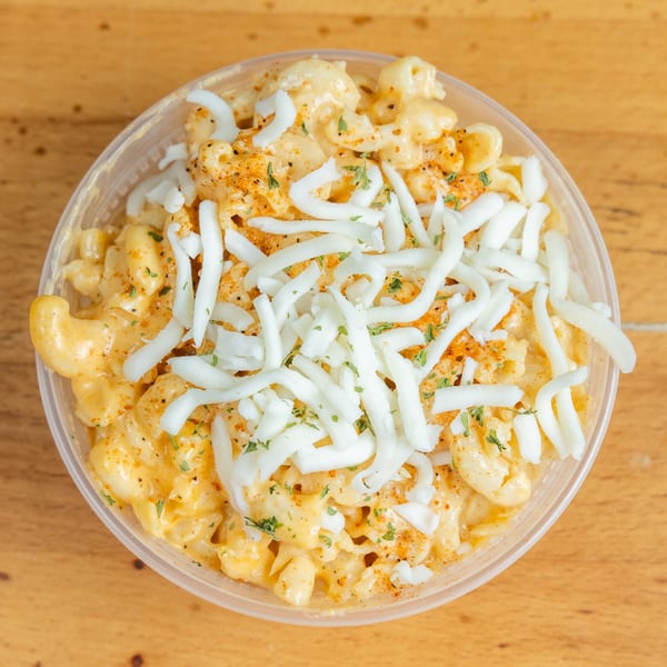 Creamy macaroni and cheese topped with shredded cheese, herbs, and spices in a clear container on a wooden table.