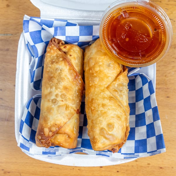 Two golden fried empanadas served in a blue-checkered basket with a cup of orange dipping sauce.